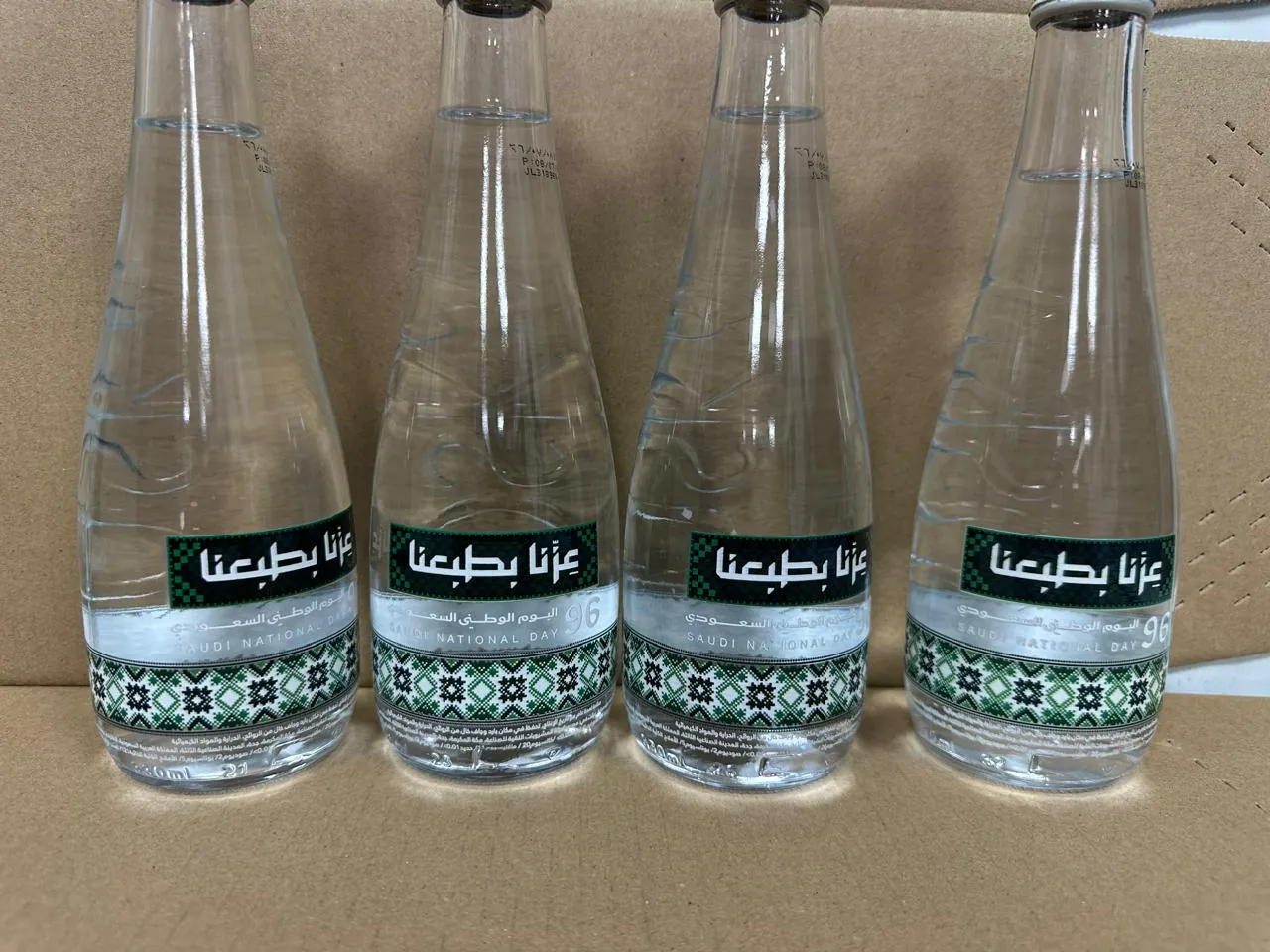 Four glass water bottles printed directly with the Saudi National Day slogan عزنا بطبعنا and a green geometric band